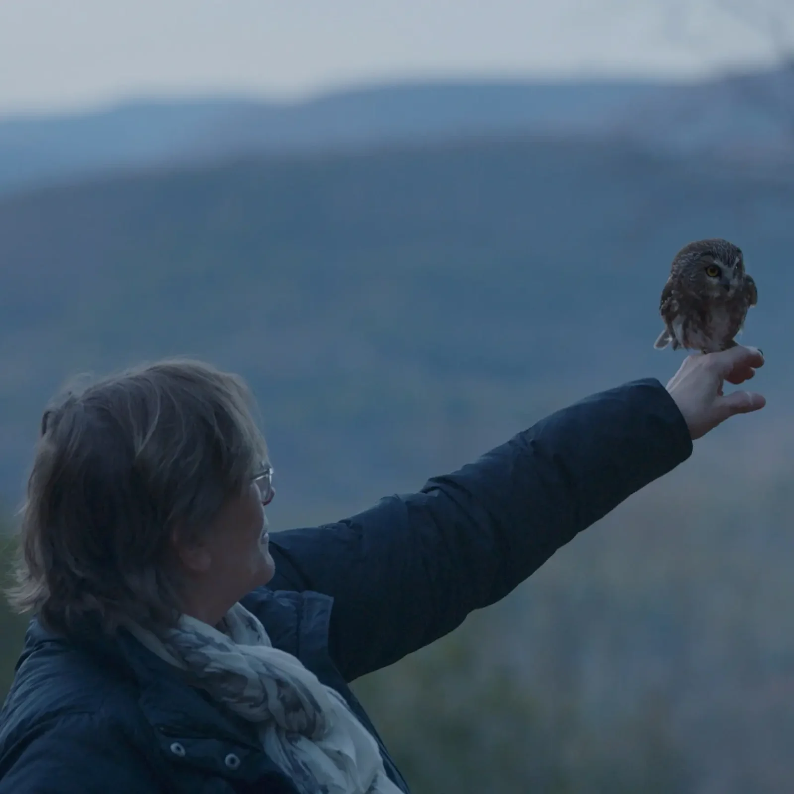 Ellen raises Rocky on an outstretched arm in a wooded outdoor scene.